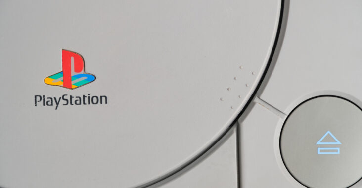 Auf dem Bild ist ein Ausschnitt einer PlayStation-Konsole zu sehen. Im Zentrum befindet sich das farbenfrohe PlayStation-Logo, das durch das charakteristische "P" und vier farbige Felder in den Formen eines Dreiecks, Kreises, Kreuzes und Quadrats dargestellt wird, welche als Richtungstasten auf dem PlayStation-Controller bekannt sind. Unter dem Logo ist der Schriftzug "PlayStation" in schwarzen Buchstaben zu erkennen. Rechts ist ein silberner, runder Knopf mit einem blauen Dreieckssymbol zu sehen, was auf eine Benutzeroberfläche oder einen Knopf auf der Konsole hindeutet. Der Hintergrund ist hellgrau, und das gesamte Design ist minimalistisch und sauber, wie es für elektronische Produkte typisch ist